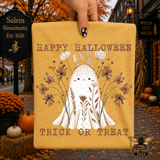 A hand holds a yellow t-shirt featuring a cute white ghost surrounded by autumn leaves and flowers, with the text 'HAPPY HALLOWEEN TRICK OR TREAT'. The scene is set outdoors in Salem, Massachusetts, with Halloween decorations, pumpkins, and fall foliage.