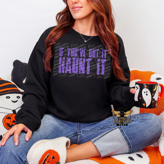 A woman wearing a black long-sleeve sweatshirt with purple text that reads 'IF YOU'VE GOT IT HAUNT IT', paired with blue jeans and white pumpkin-patterned slippers, sitting on a cozy Halloween-themed couch and holding a black mug with a ghost design.