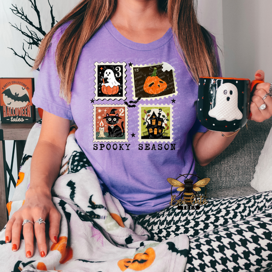 A woman wearing a purple t-shirt with a 'Spooky Season' Halloween graphic print, holding a black mug with a white ghost, while wrapped in a black and white houndstooth blanket with orange pumpkin accents.