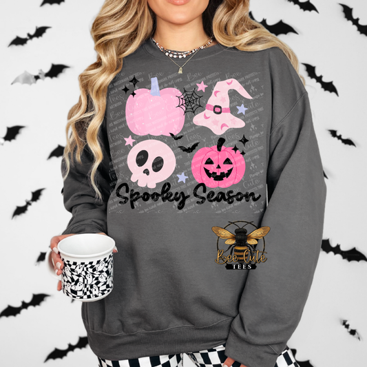 A woman wearing a dark gray sweatshirt with a pink and white 'Spooky Season' graphic print, holding a black and white patterned mug. The background is white with black bat silhouettes and features a bee logo.