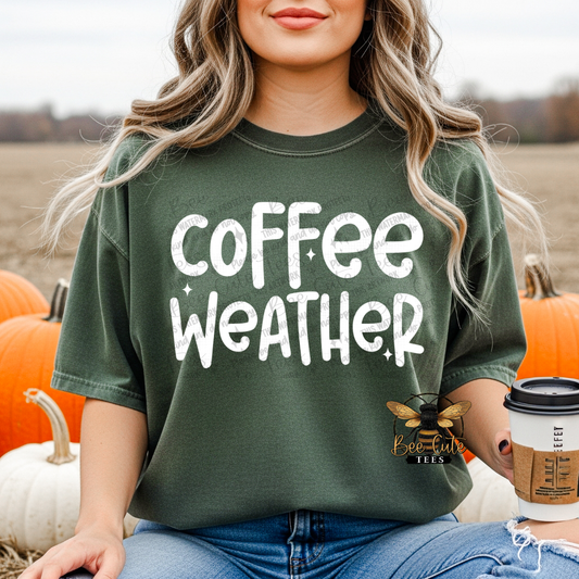 A woman wearing an oversized dark green t-shirt with 'coffee weather' printed in white distressed lettering, sitting in a pumpkin patch and holding a coffee cup.