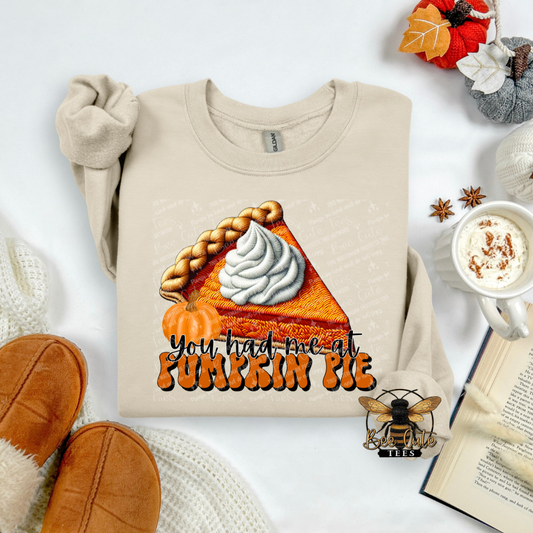 A beige long-sleeve t-shirt with a 'You had me at Pumpkin Pie' graphic, styled in a cozy flat-lay arrangement with a mug of coffee, knit socks, an open book, and autumn leaves.