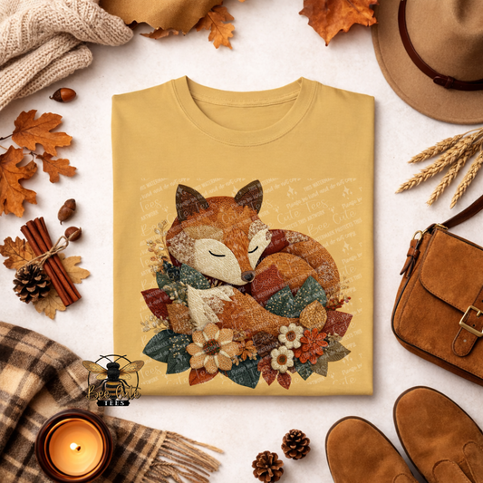 Folded mustard yellow t-shirt featuring a sleeping woodland fox surrounded by fall flowers and foliage in a vintage patchwork and faux embroidered style. Styled with autumn leaves, a cozy scarf, candle, pinecones, and rustic accessories. Bee Cute Tees watermark visible.