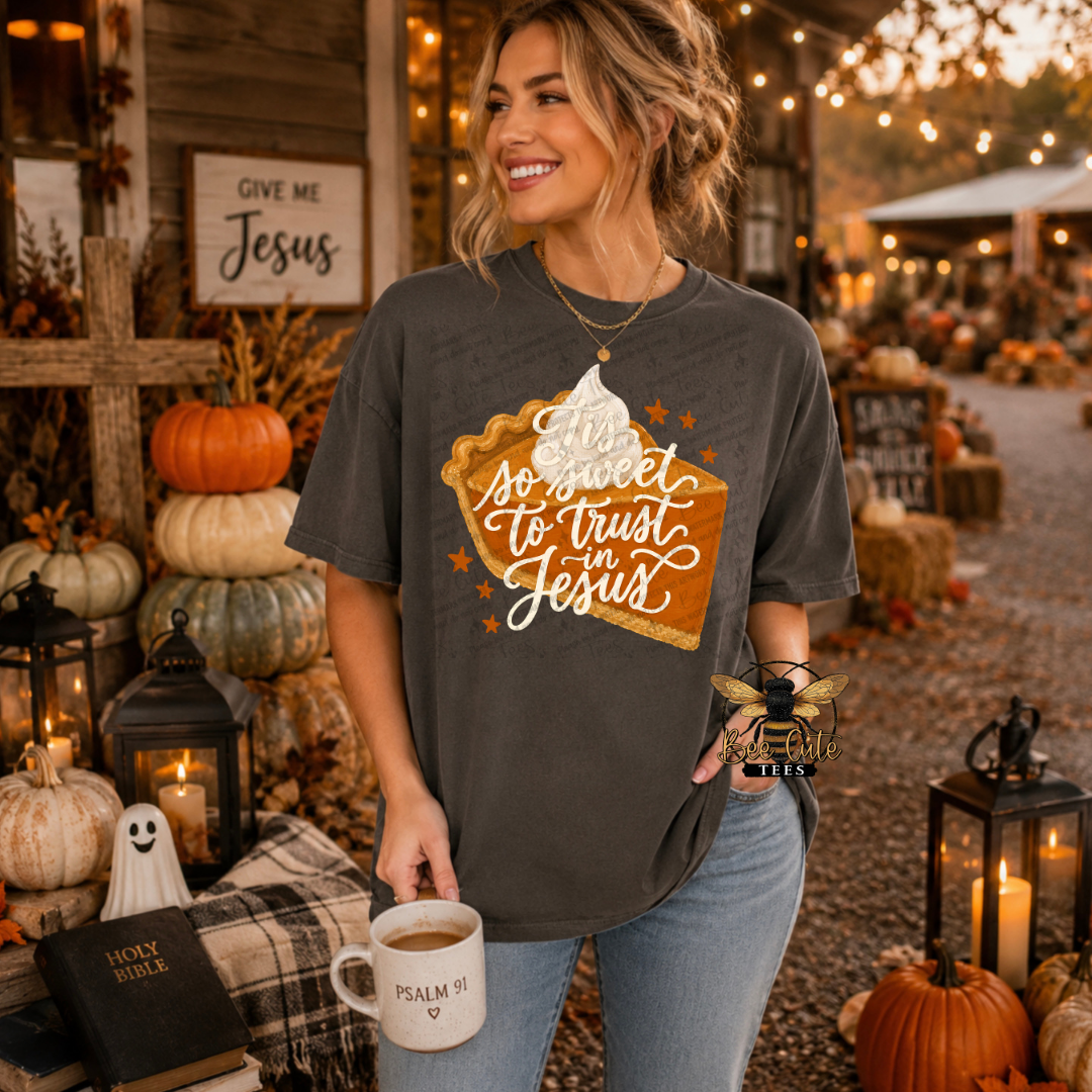 A woman stands outdoors in a fall-themed setting wearing a dark charcoal gray graphic t-shirt. The shirt features a pie graphic with whipped cream and the text 'so sweet to trust to Jesus in Jesus' in white script.