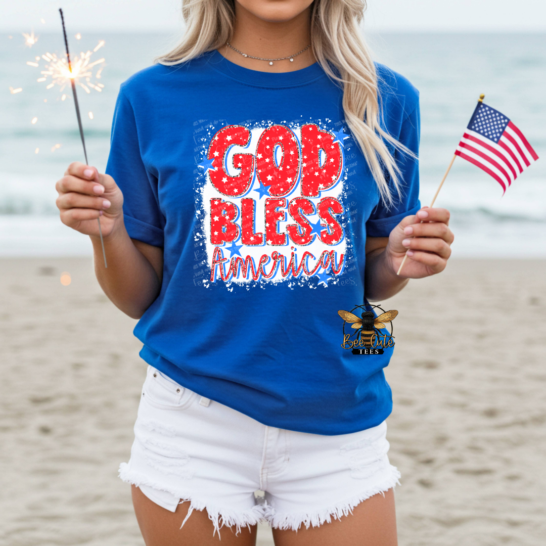 Person wearing a blue 'God Bless America' t-shirt on a beach with an American flag and sparkler.