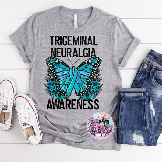 A gray short-sleeve t-shirt with 'Trigeminal Neuralgia Awareness' text and a butterfly graphic, displayed flat with sneakers and jeans on a white wooden background.
