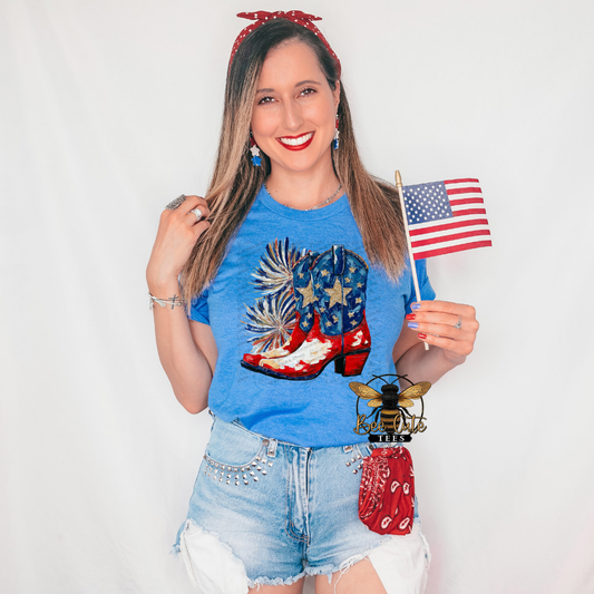 Woman wearing a heather royal blue graphic tee featuring patriotic cowboy boots decorated with stars and stripes, accented by festive fireworks. Styled with distressed denim shorts, patriotic earrings, a red bandana accessory, and holding an American flag for a fun all-American country look.