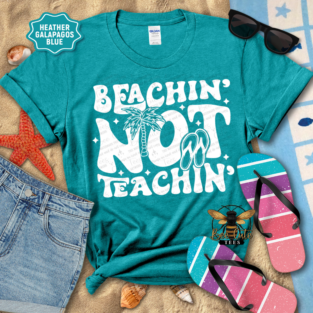 Teal t-shirt with 'Beachin' Not Teachin' design, sunglasses, flip-flops, and beach accessories on a wooden surface.