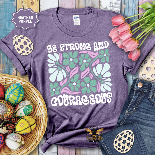 Purple t-shirt with floral design and text 'Be Strong and Courageous' on a wooden surface with Easter decorations.
