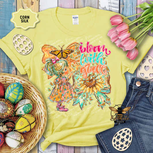 Yellow t-shirt with a colorful design featuring a fairy, butterfly, and sunflower text 'Bloom with Flare' on a wooden surface with Easter decorations.