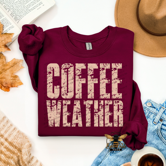 Maroon sweatshirt with 'Coffee Weather' text on a white surface with autumn leaves and a hat.