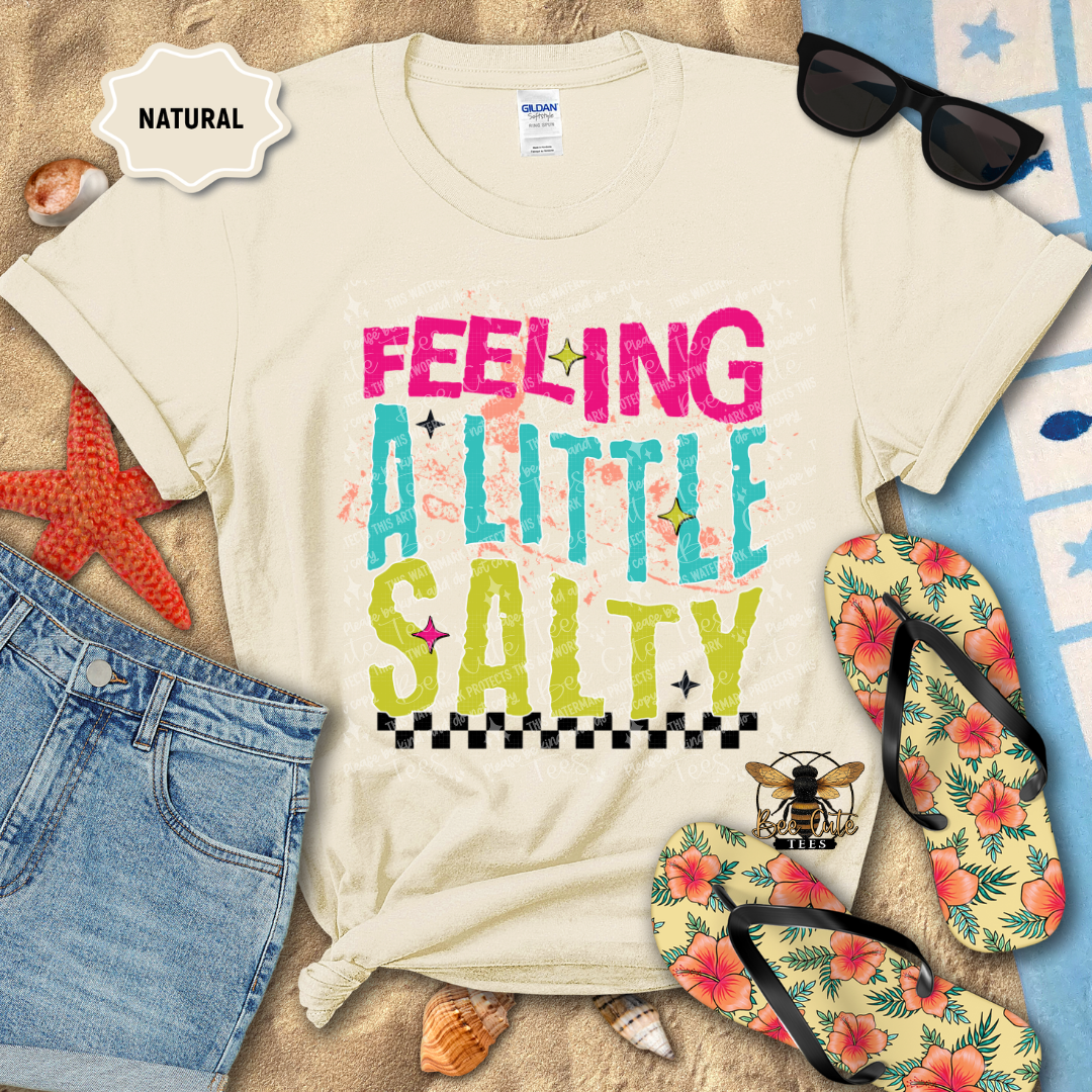 Beach-themed t-shirt with 'Feeling a Little Salty' text, sunglasses, flip-flops, and beach accessories on sand.