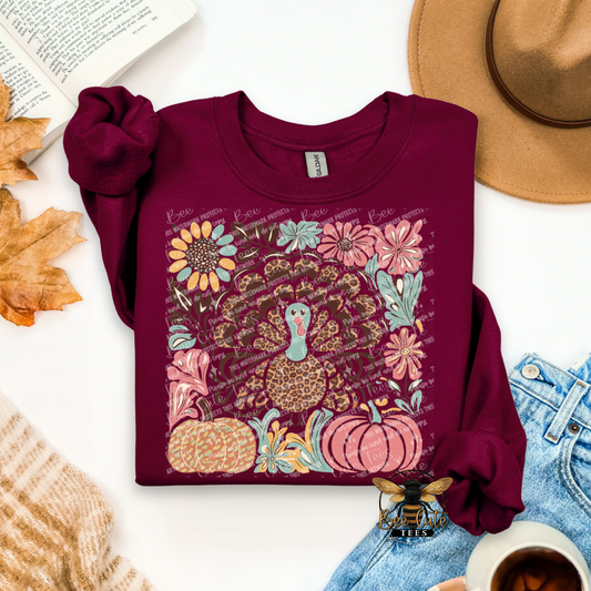 Maroon sweatshirt with a colorful turkey and floral design on a white surface with autumn leaves and a hat.