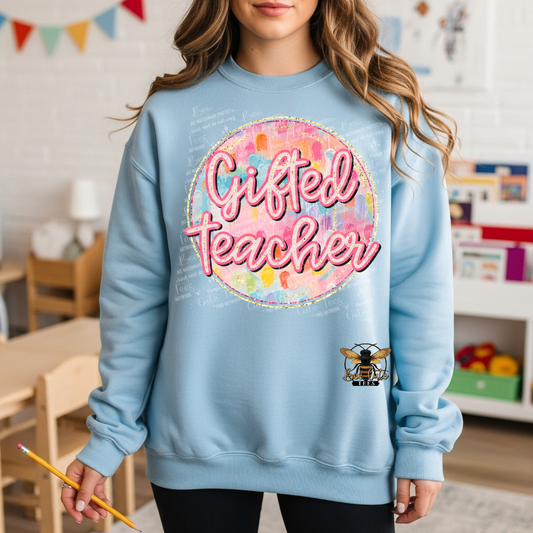 A woman wearing a light blue crewneck sweatshirt with a colorful 'Gifted Teacher' graphic and a bee logo on the front, set in a classroom environment.