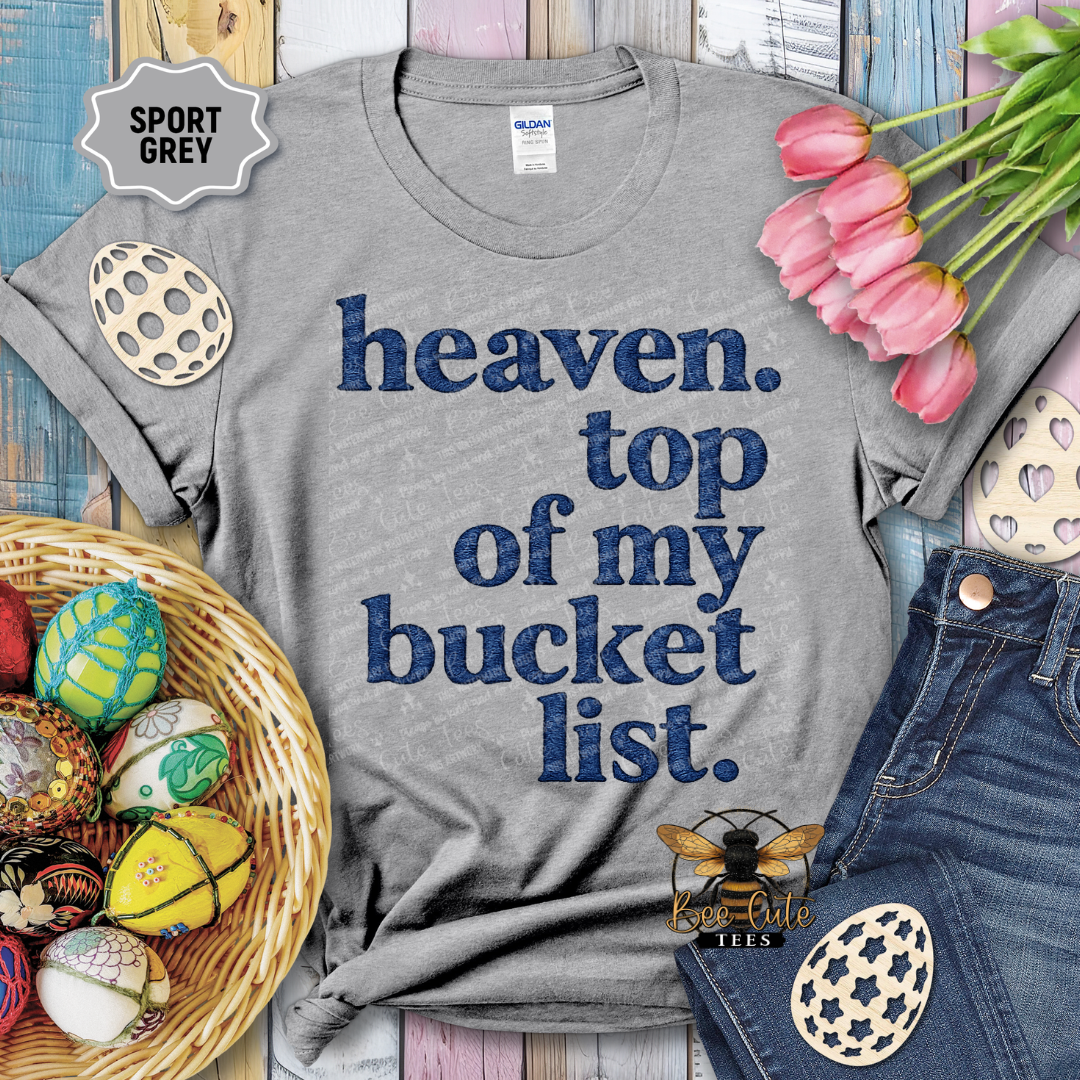 Gray t-shirt with 'heaven. top of my bucket list.' text, surrounded by Easter decorations on a wooden surface.