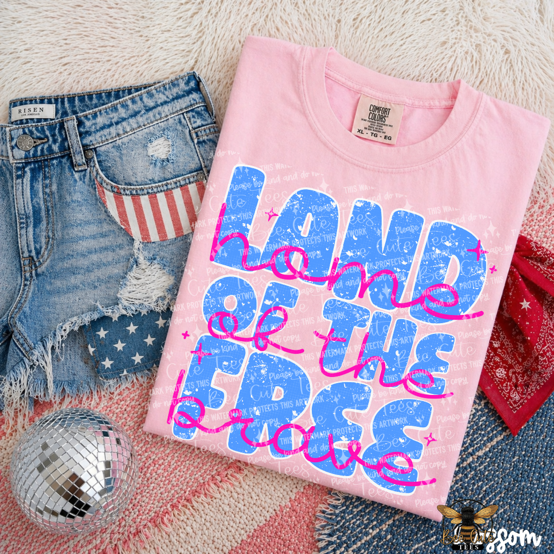 Pink t-shirt with 'Land of the Free because of the Brave' text on a textured surface with denim shorts and disco ball.