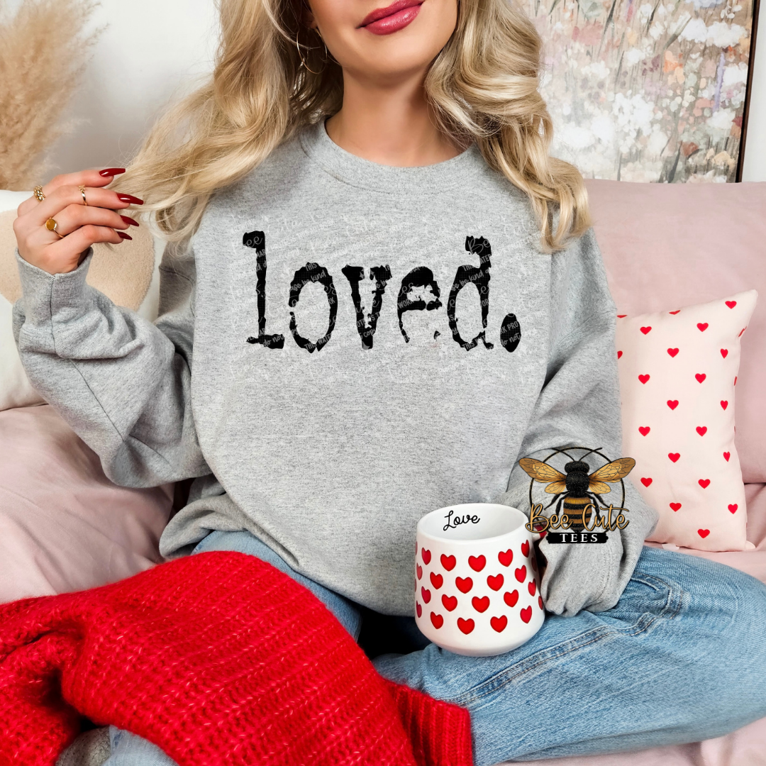 Person wearing a gray sweatshirt with 'loved.' text, sitting on a couch with a mug and decorative pillows.