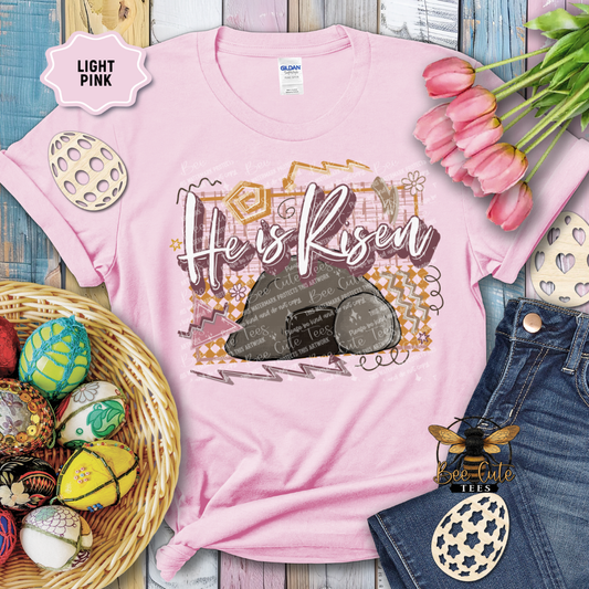 Pink t-shirt with 'He is Risen' design, surrounded by Easter decorations on a wooden surface.