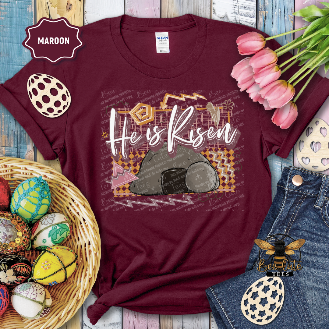 Maroon t-shirt with 'He is Risen' design, surrounded by Easter decorations on a wooden surface.
