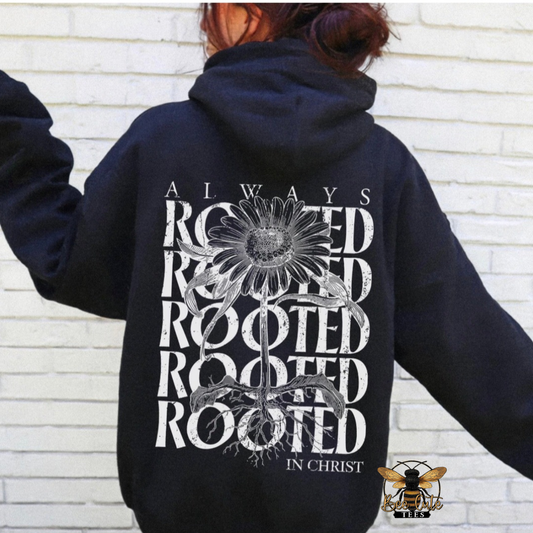 A person wearing a black hoodie from the back, featuring a large white graphic print with a sunflower and the words 'ALWAYS ROOTED IN CHRIST'. The person is standing against a white brick wall.
