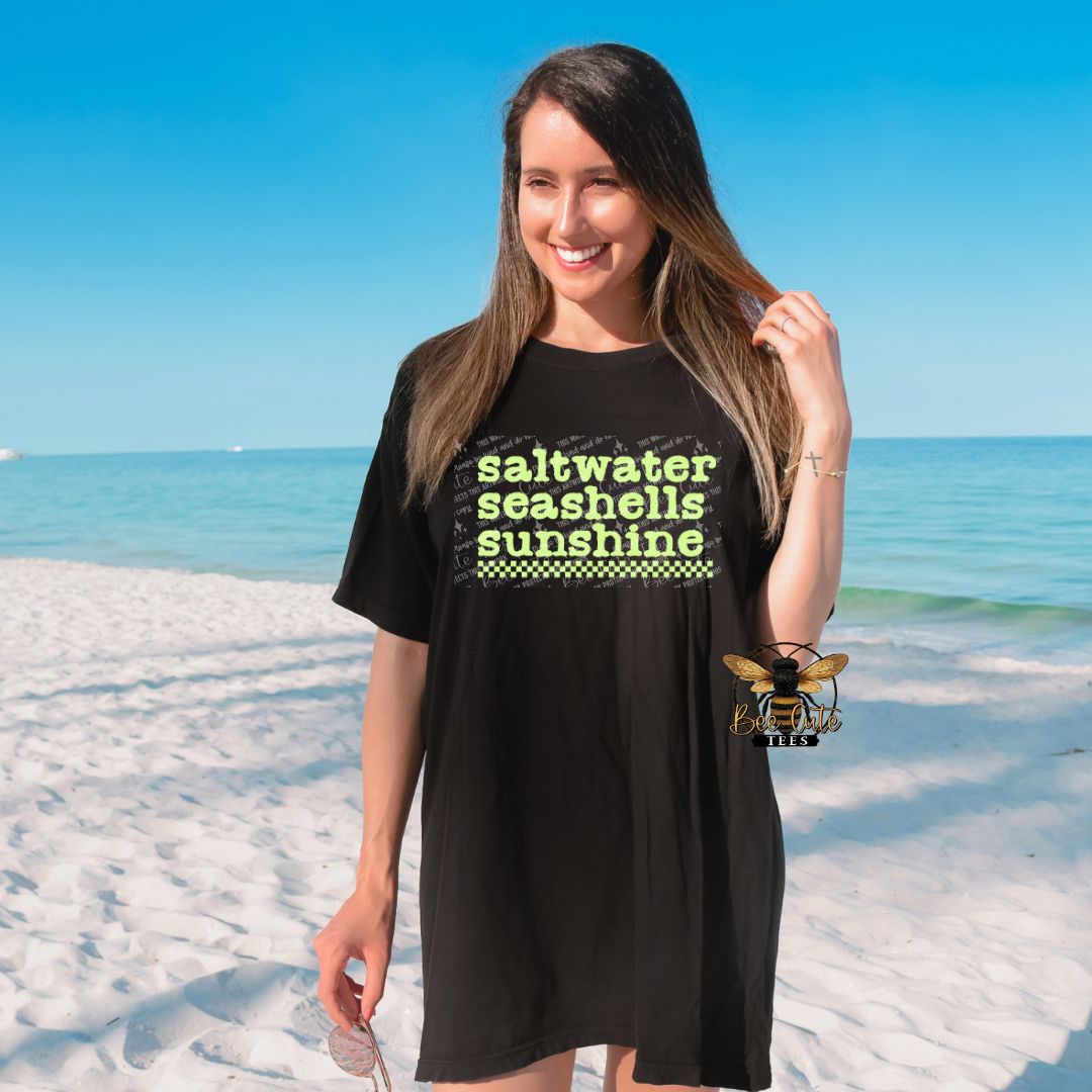 Woman wearing a black t-shirt with 'saltwater seashells sunshine' text on a beach.