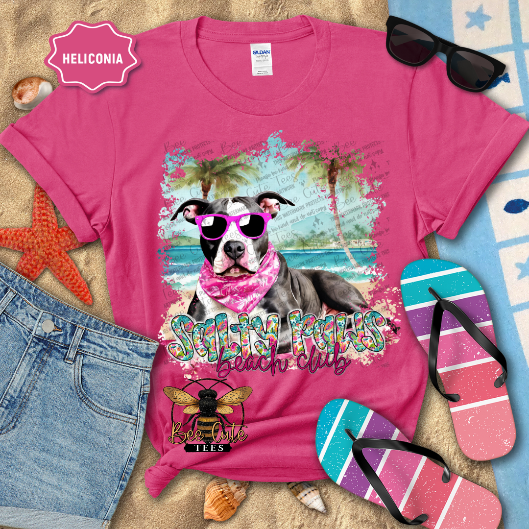 Pink t-shirt with a dog graphic and 'Salty Paws Beach Club' text, surrounded by beach accessories.