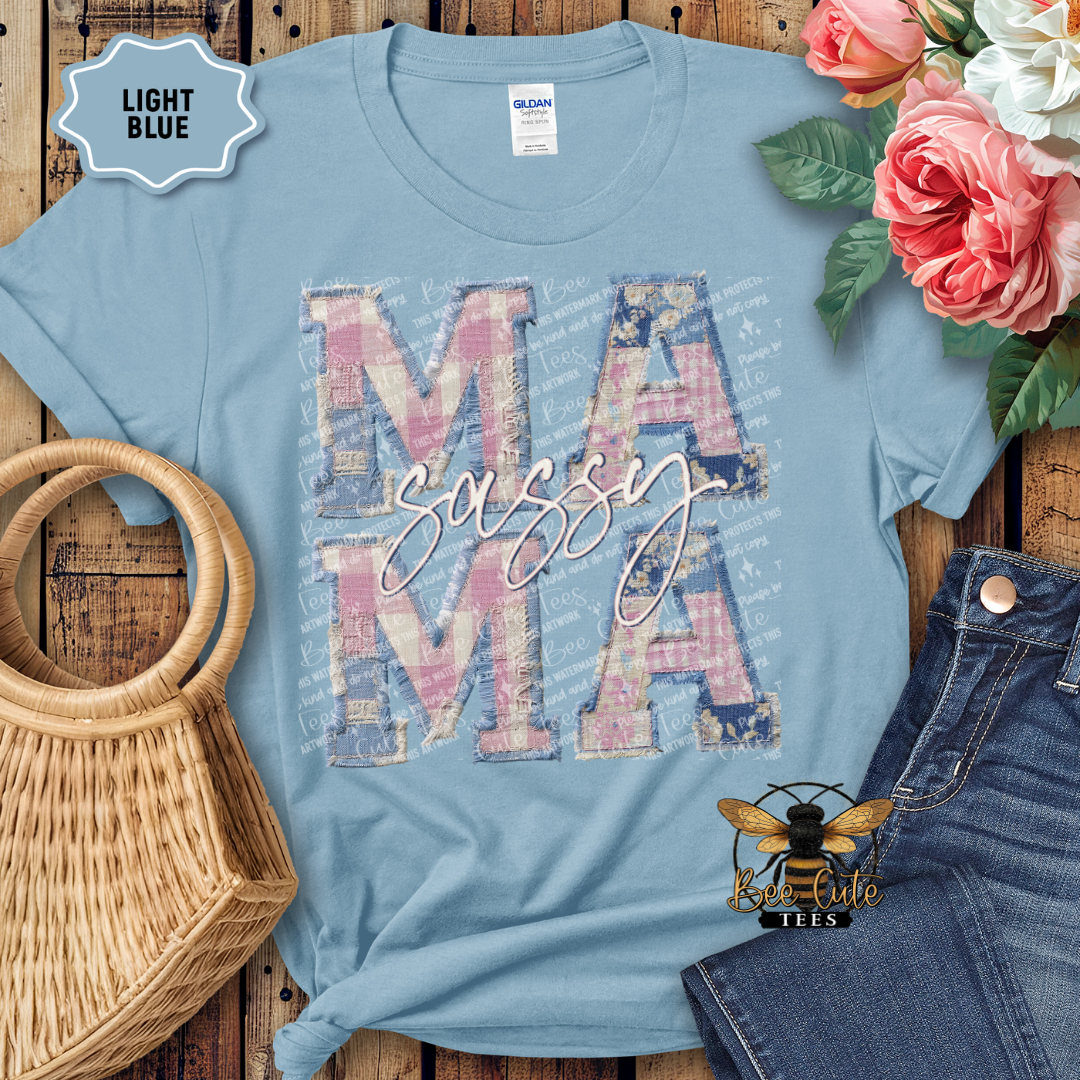 Light blue t-shirt with 'MAMA sassy' design on a wooden surface with flowers and denim jeans.
