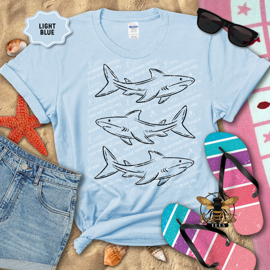 Light blue t-shirt with shark designs on a sandy background with beach accessories.