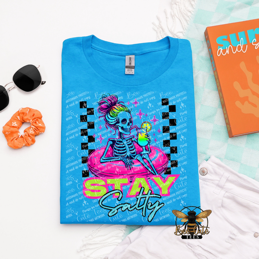 Blue t-shirt with a skeleton design and 'Stay Salty' text, surrounded by sunglasses, scrunchie, and notebook.