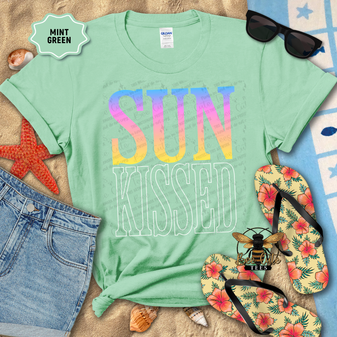 Mint green t-shirt with 'SUN KISSED' text, sunglasses, flip-flops, and beach accessories on a sandy background.