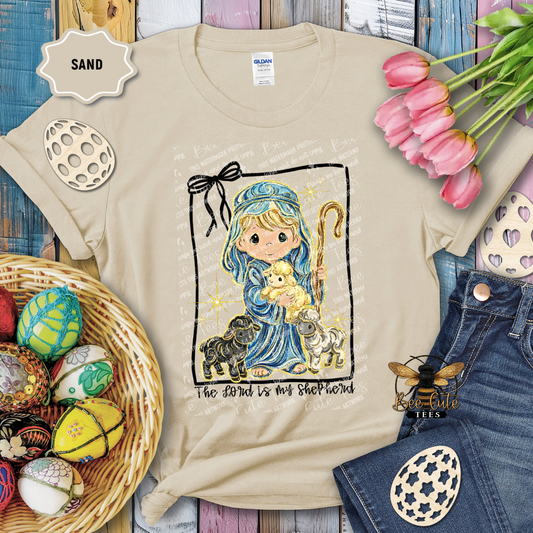 Beige t-shirt with a graphic design of a shepherd holding a lamb, surrounded by Easter decorations.