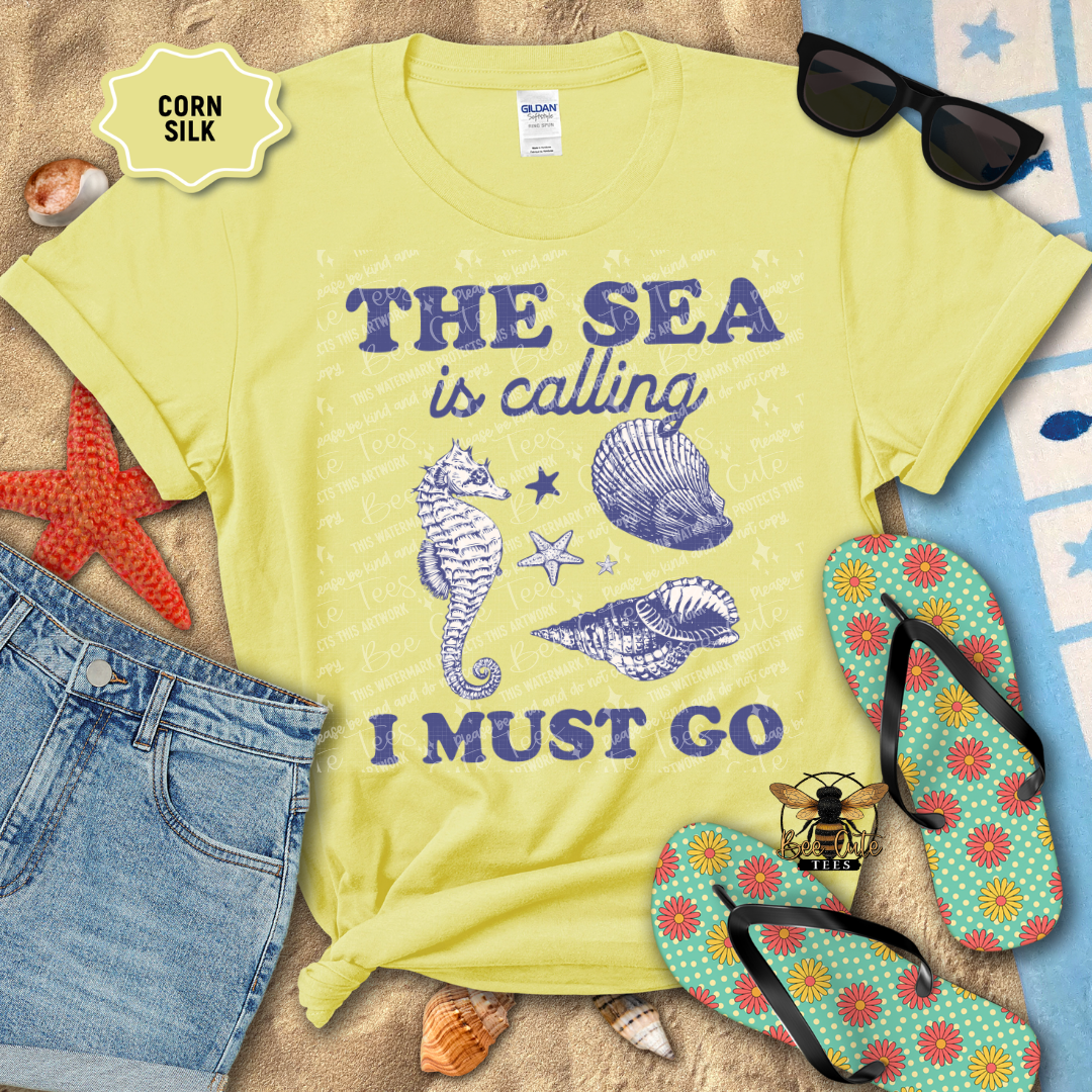 Yellow t-shirt with 'The Sea is Calling I Must Go' text and beach-themed graphics on a sandy background.