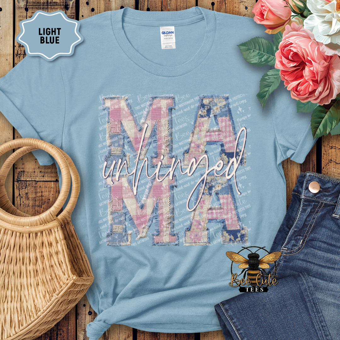 Light blue t-shirt with 'MAMA unHINGED' design on a wooden surface with flowers and jeans.