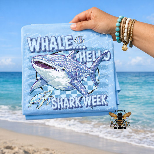A folded light blue T-shirt is held up by a person's hand against a beach backdrop. The shirt features a large whale shark graphic with the text 'WHALE.