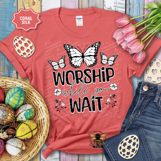 Red t-shirt with butterfly design and text 'Worship while you wait' on a wooden surface with Easter decorations.
