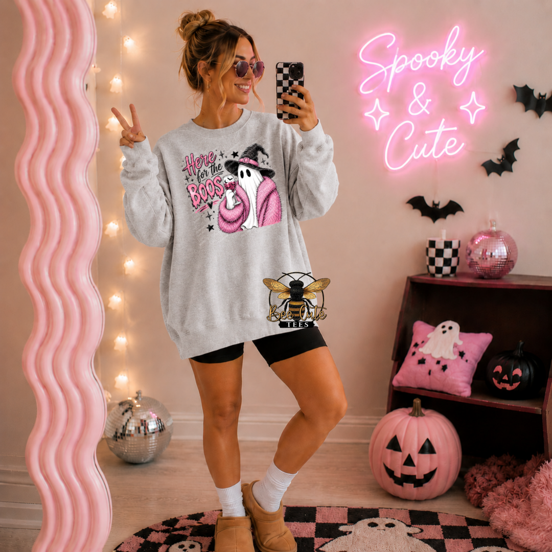 A woman takes a mirror selfie wearing a light gray crewneck sweatshirt with a pink and black witch ghost graphic and the text 'Here For The Boos'. She is posing in a decorated room with pink and black Halloween decor, including a pink wavy mirror, a pink neon sign that reads 'Spooky & Cute', pumpkins, and a shelf with ghost and pumpkin pillows.