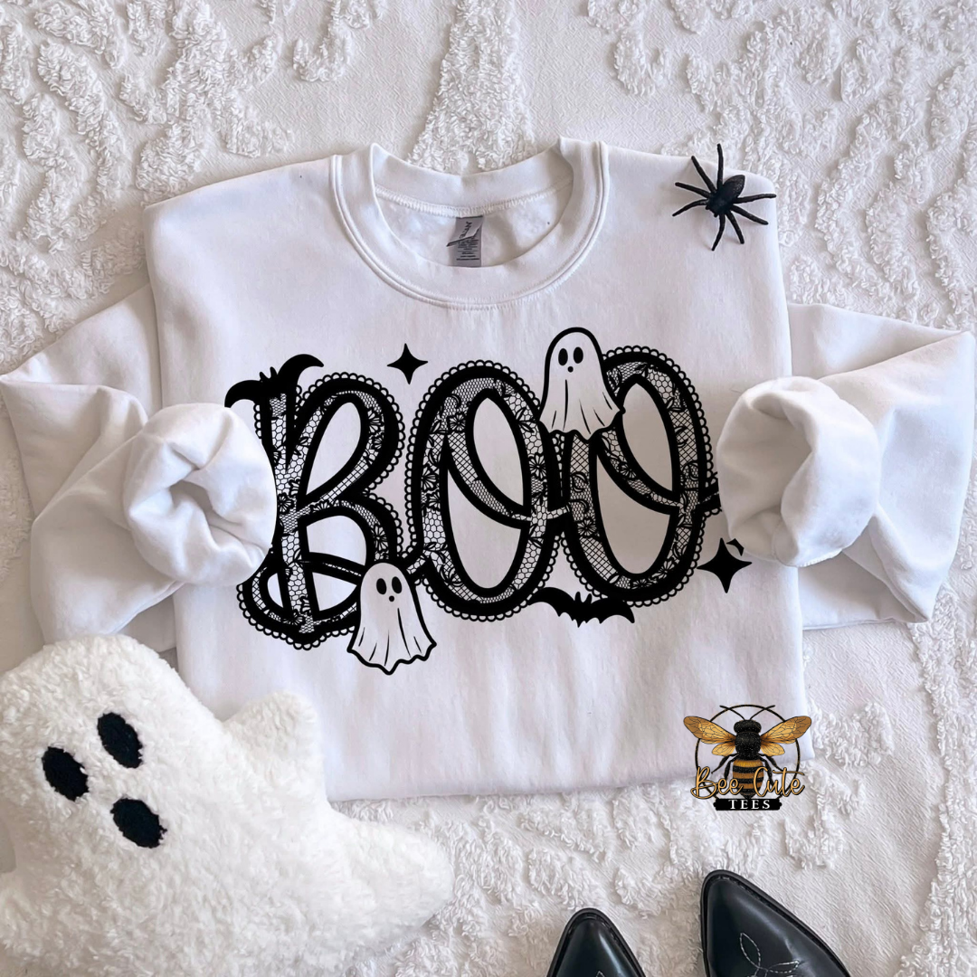 A white long-sleeve sweatshirt with a black 'BOO' Halloween graphic featuring ghosts, bats, and stars, styled in a flat lay on a textured white blanket with a plush ghost, a spider decoration, and black shoes.