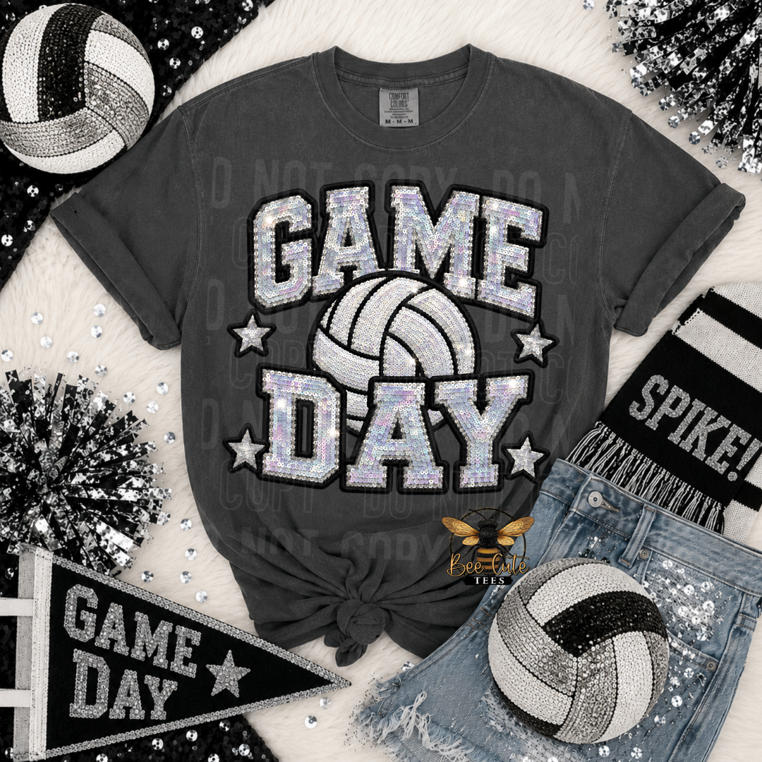 A dark gray graphic t-shirt featuring a 'GAME DAY' volleyball design in a glittery silver and white pattern, styled in a flat-lay with denim shorts, a volleyball, a scarf, and decorative tinsel.