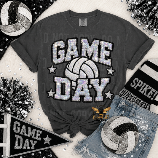 A dark gray graphic t-shirt featuring a 'GAME DAY' volleyball design in a glittery silver and white pattern, styled in a flat-lay with denim shorts, a volleyball, a scarf, and decorative tinsel.