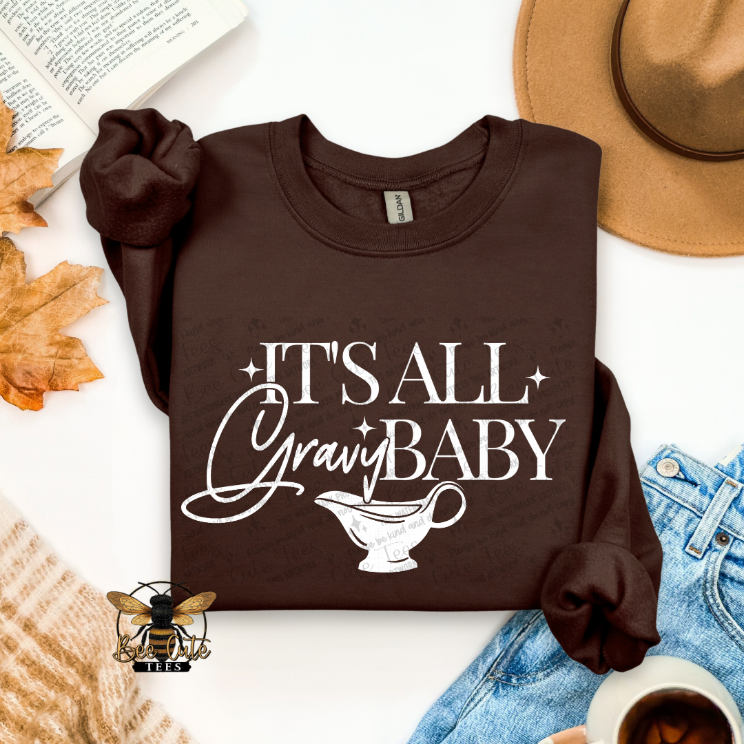A dark brown long-sleeve t-shirt with a white graphic reading 'IT'S ALL Gravy BABY' and a gravy boat illustration, styled in a flat lay with a hat, jeans, leaves, a book, and a cup on a white background.