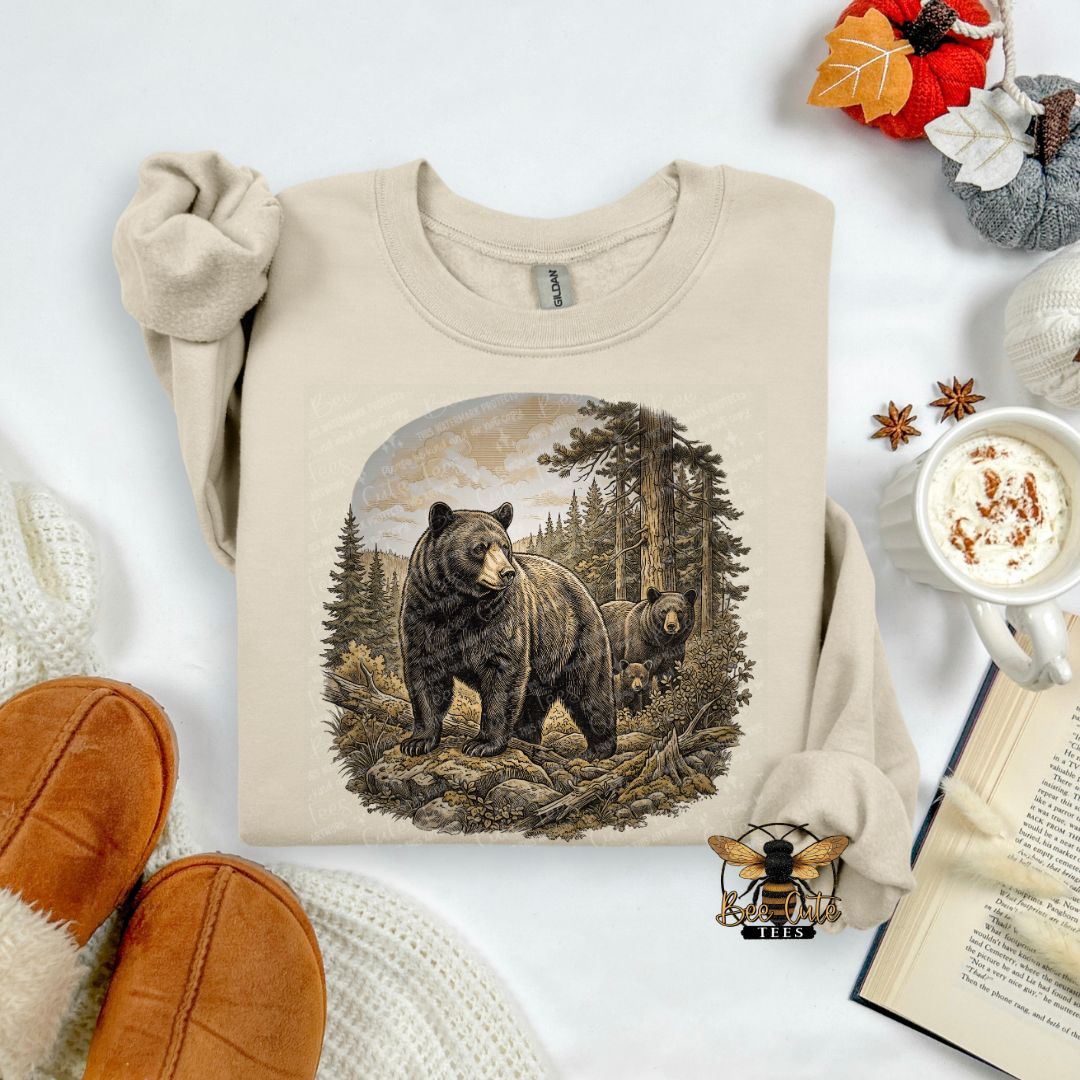 A beige crewneck sweatshirt with a detailed graphic of a black bear and cub in a forest, styled in a cozy flat-lay with brown slippers, a mug of coffee, an open book, and autumn leaves.