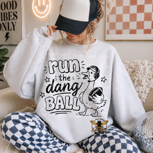 A woman sitting on a couch wearing a light gray sweatshirt with a black graphic that reads 'Run the dang ball' and shows determination with a football. She is also wearing a black and white trucker hat and blue and white checkered pajama pants.