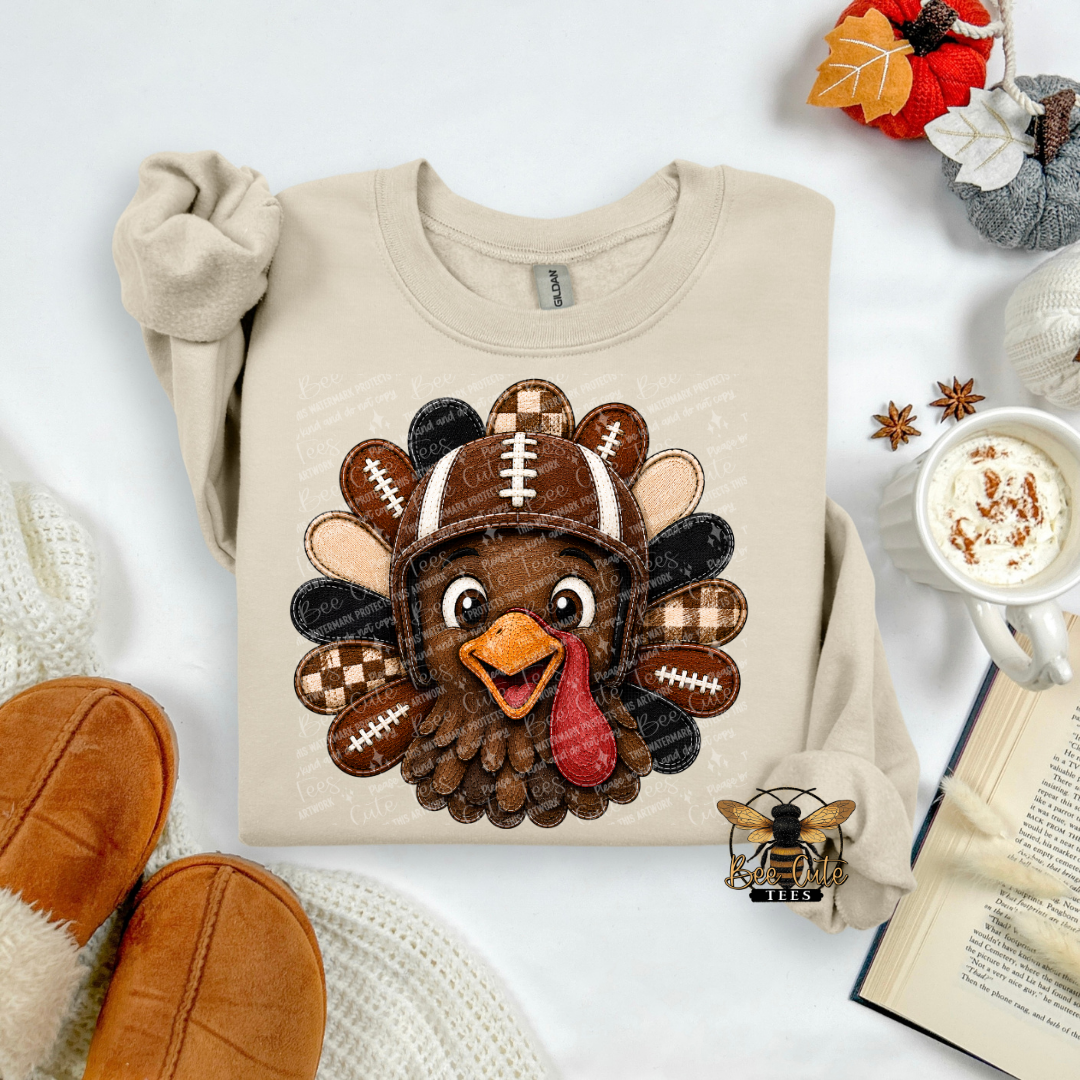 A beige long-sleeve sweatshirt laid flat on a white surface, featuring a large cartoon turkey graphic with a football helmet. The sweatshirt is styled with fall props including a mug of hot cocoa, an open book, a knit blanket, and brown slippers.
