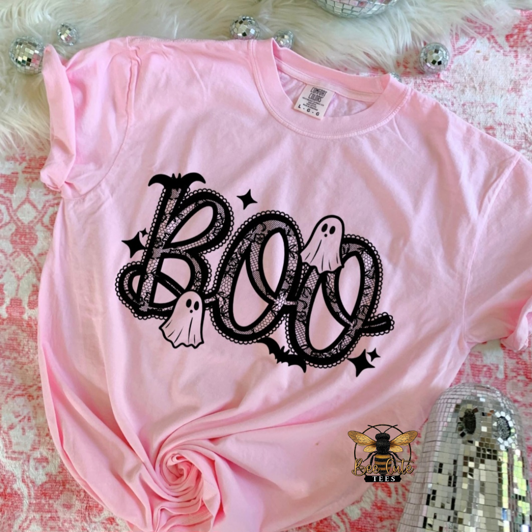 A light pink short-sleeve t-shirt with a black 'BOO' Halloween graphic featuring bats, ghosts, and stars, styled on a pink and white patterned surface with a white faux fur background.