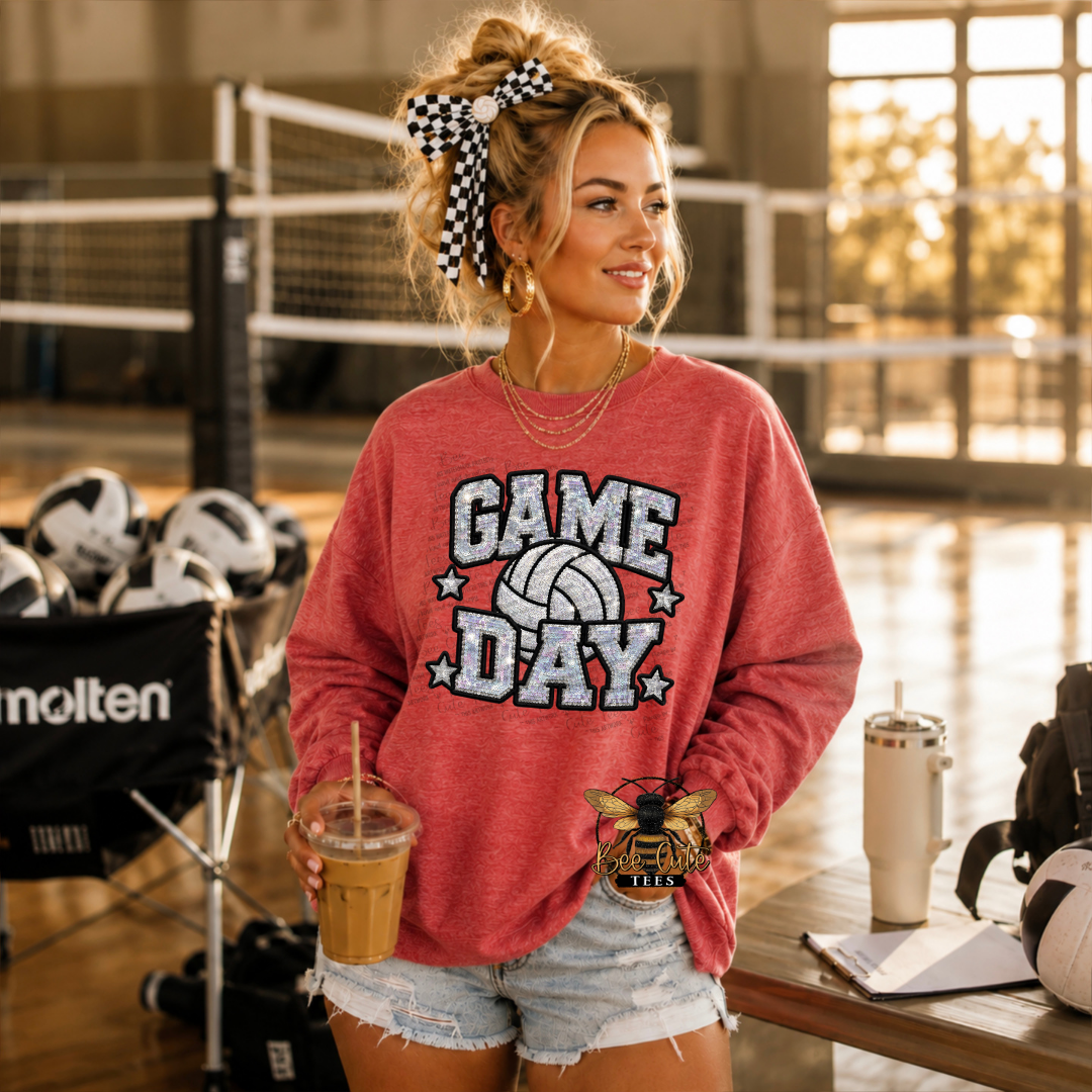 A woman stands in a volleyball gym wearing a heathered red graphic sweatshirt that says 'GAME DAY' with a volleyball and bee logo, paired with denim shorts.
