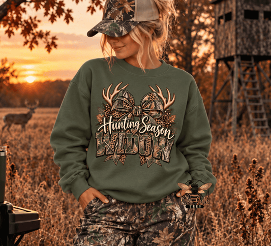 A woman wearing an olive green sweatshirt with a camouflage hunting-themed graphic that reads 'Hunting Season Widow' and antlers, standing in a field at sunset with deer and a camouflage hunting stand in the background.