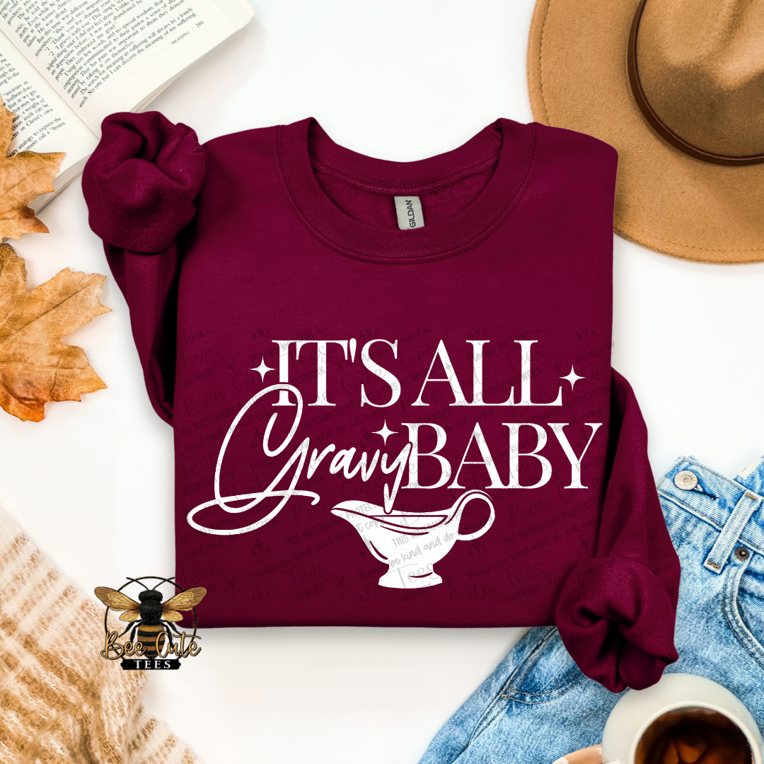 A folded maroon long-sleeve T-shirt with rolled cuffs, featuring the white text 'IT'S ALL Gravy BABY' and a gravy boat graphic on the front. The shirt is styled in a flat lay with a tan hat, blue jeans, a book, autumn leaves, a cup of coffee, and a small bee logo.