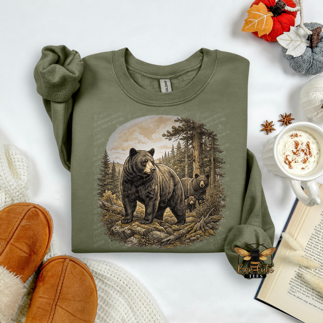 An olive green long-sleeve t-shirt with a detailed graphic of a black bear and cub in a forest, styled in a cozy flat-lay with a mug of coffee, an open book, and brown slippers.