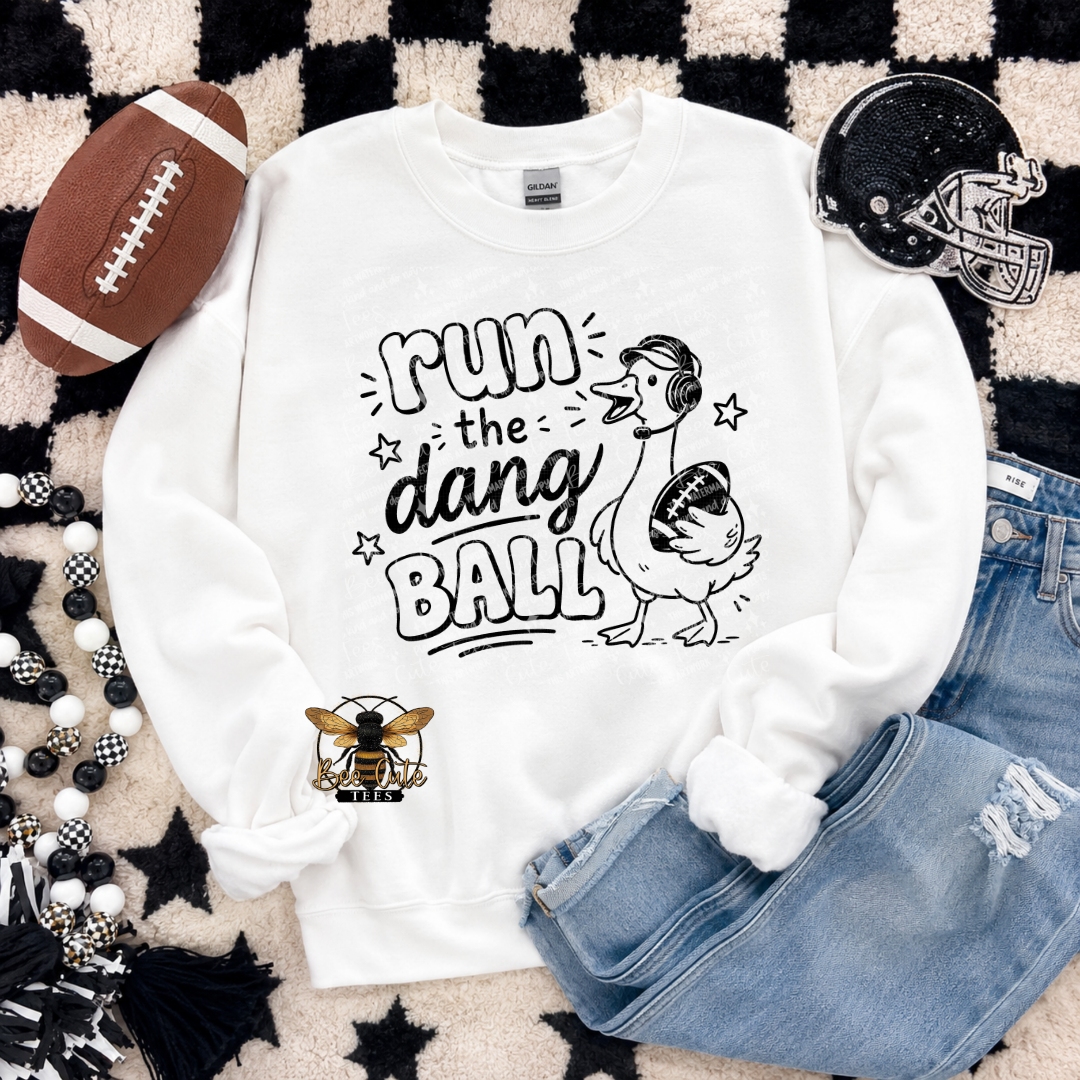 A white crewneck sweatshirt laid flat on a black-and-white checkered rug with styled props, including a football, a helmet, jeans, beads, and a bee patch graphic. The sweatshirt has a black hand-drawn-style graphic reading “run the dang ball” with a duck holding a football and a bee logo near the lower left front.