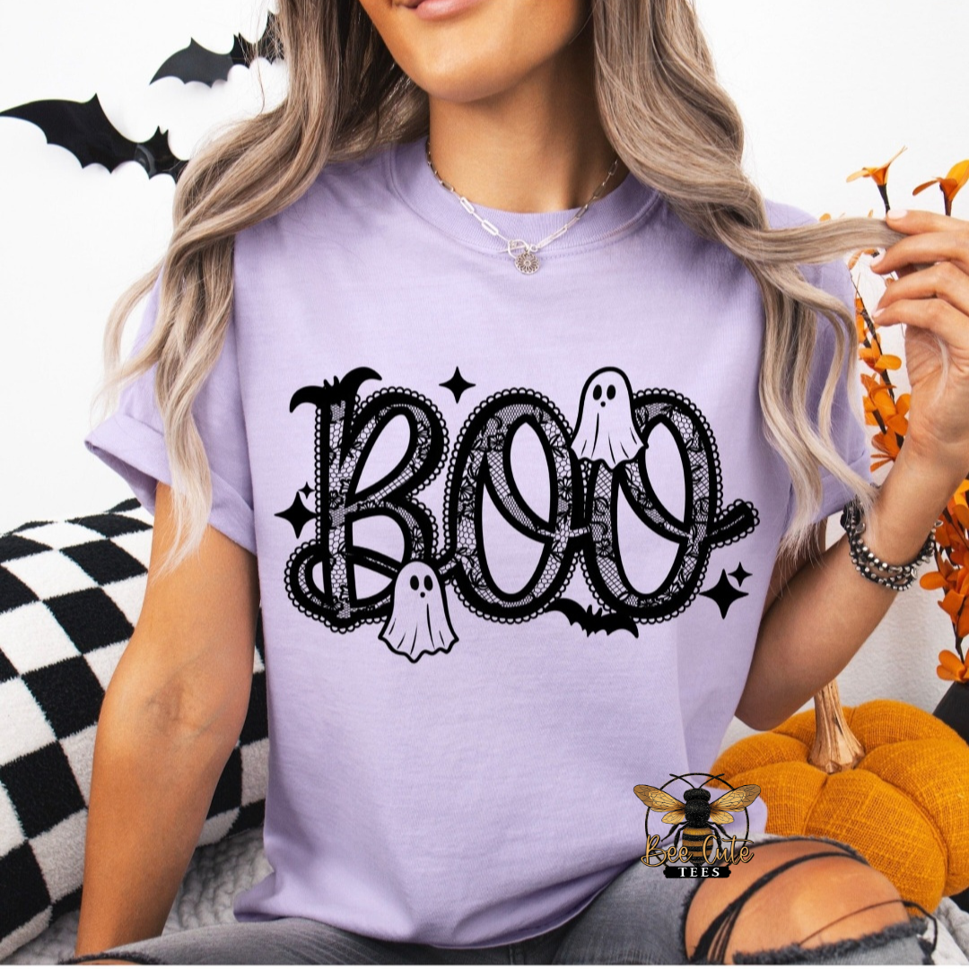 A woman wearing a light purple t-shirt with a black 'BOO' Halloween graphic featuring ghosts and bats, posed with a pumpkin and checkered pillow.